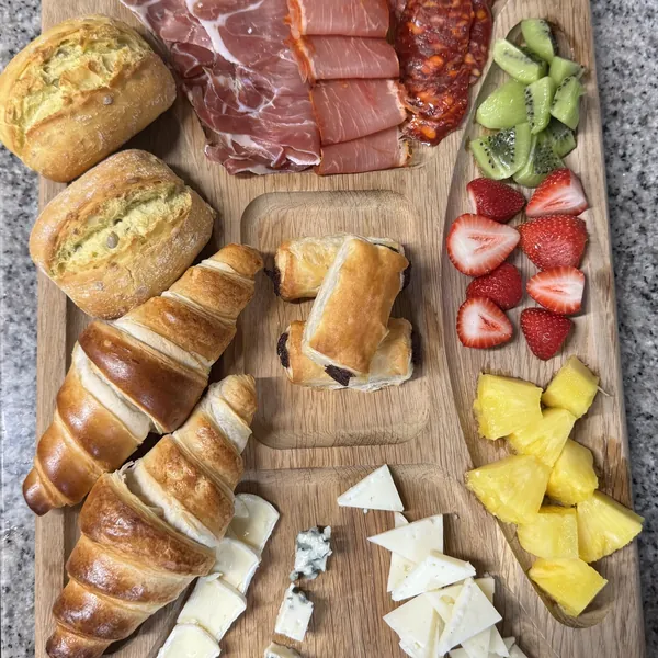 Tabla de desayuno con pan, cruasanes, jamón, chorizo, quesos y fruta